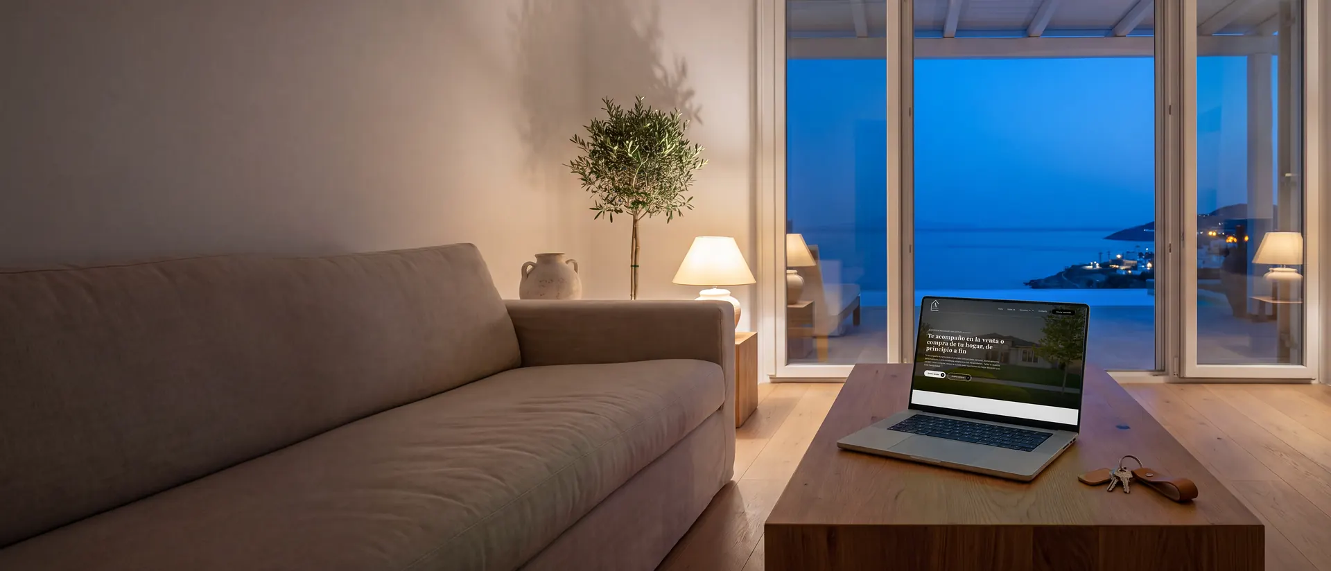 Salón de una vivienda junto al mar a la hora azul, con un portátil que muestra la web de Abad Realty, diseñada por Pc Emprende.